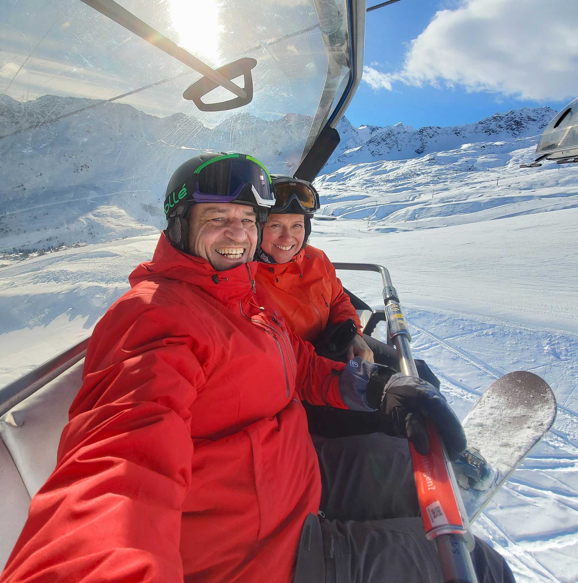 ski-und-schule-bayrischzell-staatlich-tablet Zwei lachende Wintersportler in roten Skijacken sitzen auf einem Sessellift in einem sonnigen Skigebiet mit verschneiten Bergen im Hintergrund.
