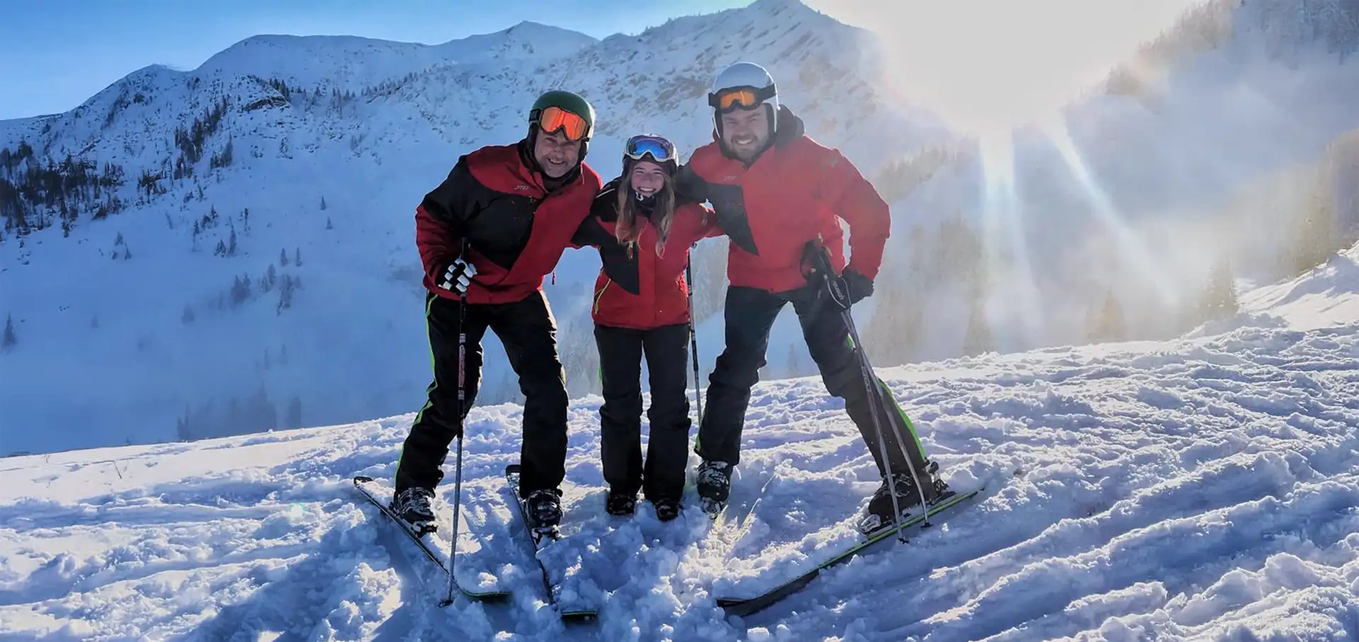 ski-und-schule-bayrischzell-team-mobil Drei Skifahrer der Skischule und Snowboardschule Bayrischzell posieren lachend auf einer sonnigen Skipiste vor verschneiter Alpenkulisse.<br />