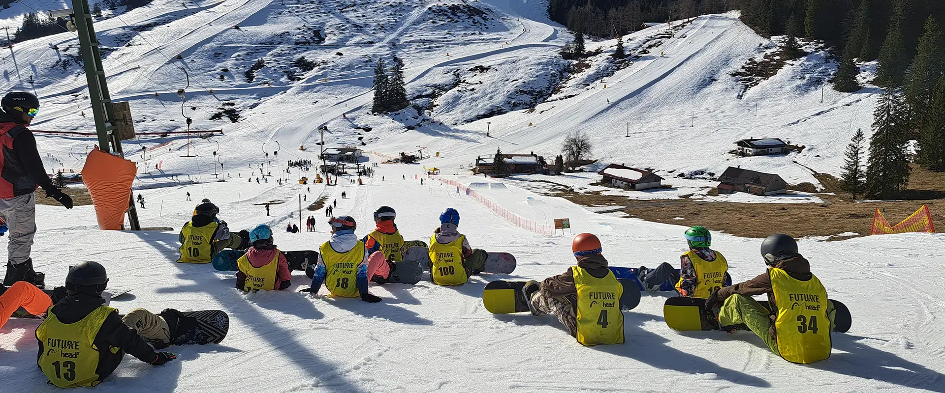 skischule_snowboardschule_bayrischzell_slide2_mobil2 Kindergruppe der Snowboardschule Bayrischzell sitzt mit Snowboards auf der Piste und bereitet sich auf die Abfahrt vor – Blick ins Tal bei sonnigem Winterwetter.